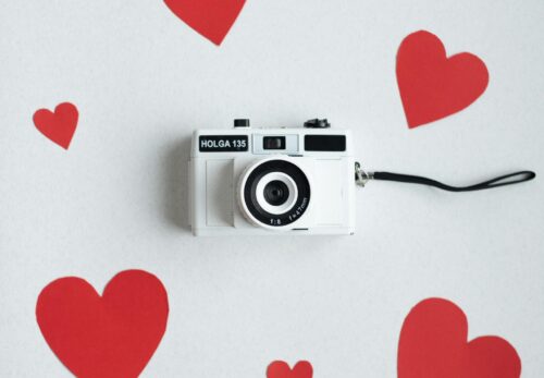 White vintage camera surrounded by red heart cutouts on a white background for a Valentine's Day theme.